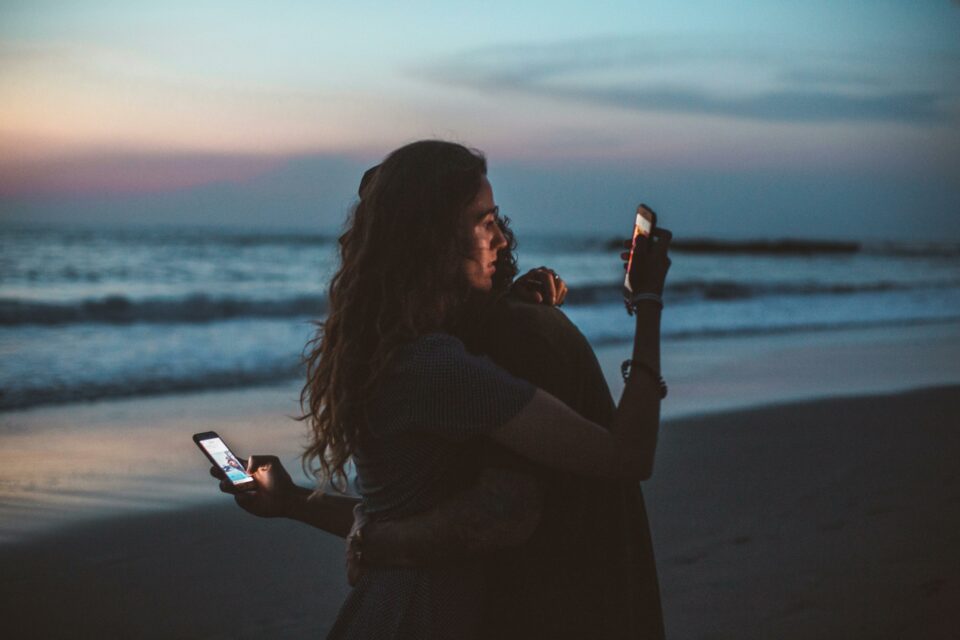 una pareja se da un abrazo en la playa con los móviles en las manos